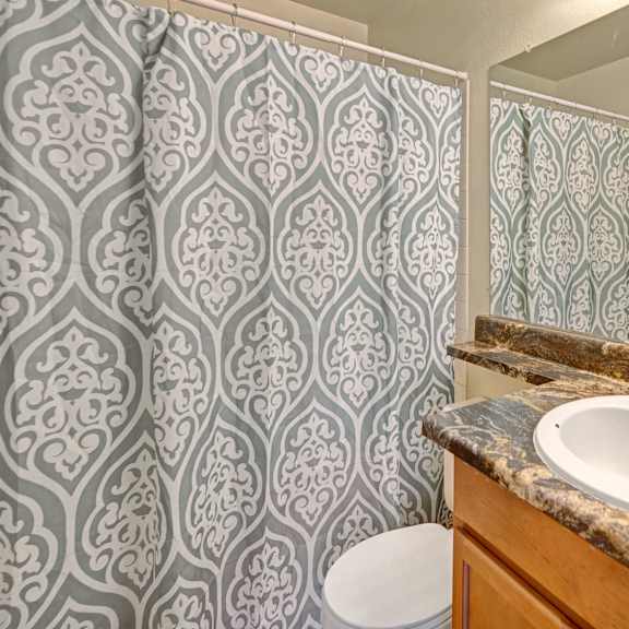 bathroom here at conifer grove apartments with a decorative green and white patterned shower curtain, a brown marble countertop, green potted plants, and a white towel on a rack.