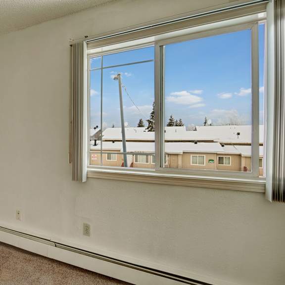 small, empty bedroom here at conifer grove apartments with beige carpet and white walls. a large window shows snowy rooftops and a clear blue sky, conveying a calm winter day.