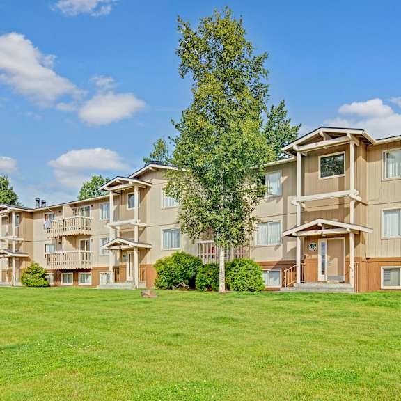 three-story beige conifer grove apartment building with white trim, surrounded by lush green grass and trees. bright blue sky with scattered clouds, conveying a serene suburban feel.