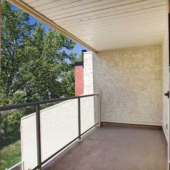covered balcony here at 3600 brentwood apartment homes with textured white walls, glass railing, and view of lush green trees against blue sky. the open door adds an inviting feel.