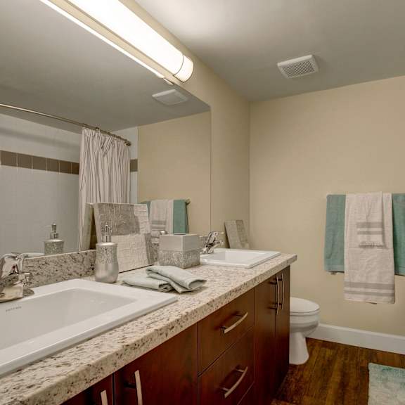 modern bathroom here at 3600 brentwood apartment homes with dual sinks on a granite countertop, wood cabinets, a shower with a striped curtain, and soft beige walls. calm and sleek ambiance.