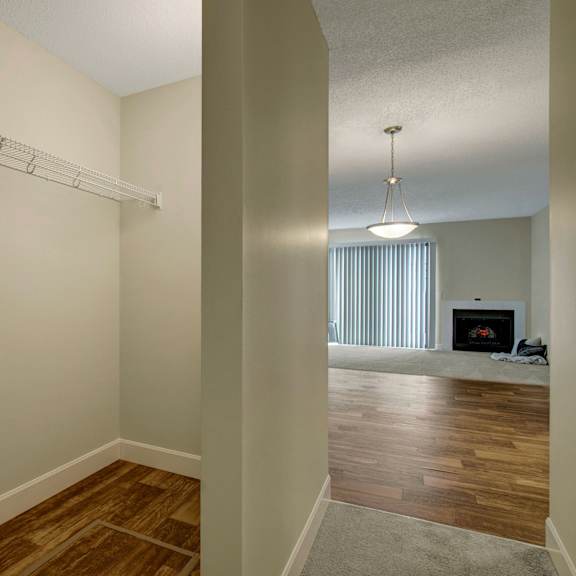 a spacious, empty walk-in closet here at 3600 brentwood apartment homes with a single wire shelf. adjacent room features wooden flooring, a lit pendant lamp, vertical blinds, and a cozy fireplace.