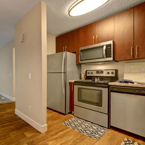 modern kitchen and living area here at 3600 brentwood apartment homes; kitchen with stainless steel appliances and wooden cabinets, opening to a living space with carpet, fireplace, and vertical blinds. cozy atmosphere.