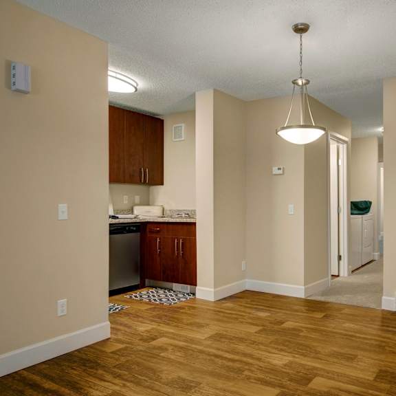 a cozy apartment here at 3600 brentwood apartment homes with wooden floors and soft beige walls. a small kitchen appears on the left with dark wood cabinets. a hallway leads to other rooms, and a crate with a "home" sign sits on the floor. a pendant light hangs from the ceiling, adding warmth.