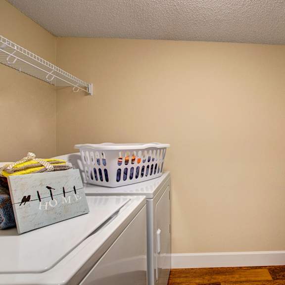 laundry room here at 3600 brentwood apartment homes with beige walls and wooden floor. a white washing machine holds a laundry basket and a "home" sign with towels on top. wire shelves line the walls.