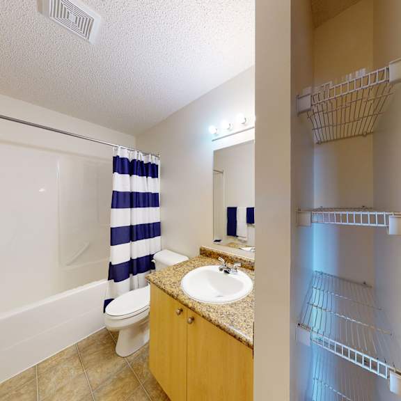 a clean bathroom here at birch terrace apartment homes with a striped blue and white shower curtain, white bathtub, and towels. a granite-top vanity with a white sink and a small mirror. an open closet with wire shelves, and a tiled floor enhancing the tidy ambiance.
