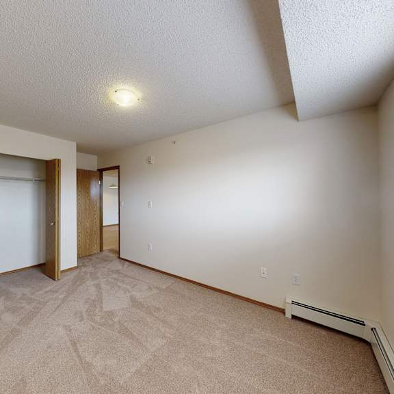 empty bedroom here at birch terrace apartment homes with beige carpet, open wooden closet doors, white walls, and a small window with blinds on the right. bright, calm atmosphere.