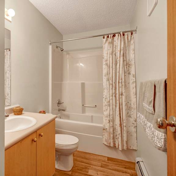 a small, cozy bathroom here at birch terrace apartment homes featuring a white sink with a wooden cabinet, a toilet, and a bathtub with a floral shower curtain. soft lighting creates a warm ambiance.