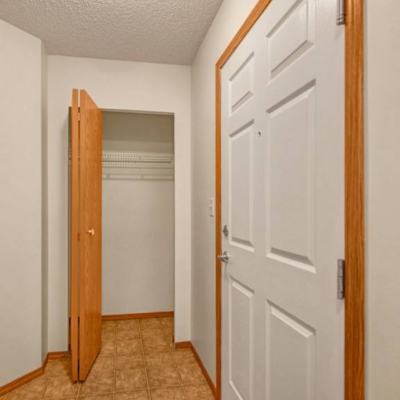 white hallway here at birch terrace apartment homes with a brown closet door ajar, revealing an empty closet with wire shelving inside. beige tile flooring and smooth pale walls. calm ambiance.