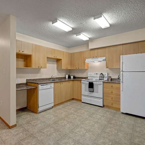 modern kitchen here at birch terrace apartment homes with light wood cabinets, granite countertops, white appliances including a fridge and stove, and overhead fluorescent lighting. neutral tones.