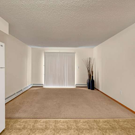 empty room here at birch terrace apartment homes with beige walls, carpet flooring, and vertical blinds covering a window. a white fridge is on the left, and tall vases with sticks are on the right.