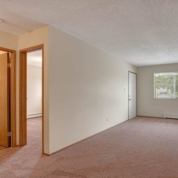 empty room here at birch terrace apartment homes with beige carpet and walls, featuring a white door, a large window with blinds, a round ceiling light, and two open doorways. cozy and minimalistic.