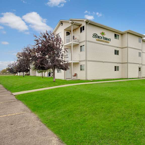 three-story white building labeled "birch terrace" amid vibrant green lawns and trees under a clear blue sky, evoking calm and suburban living.