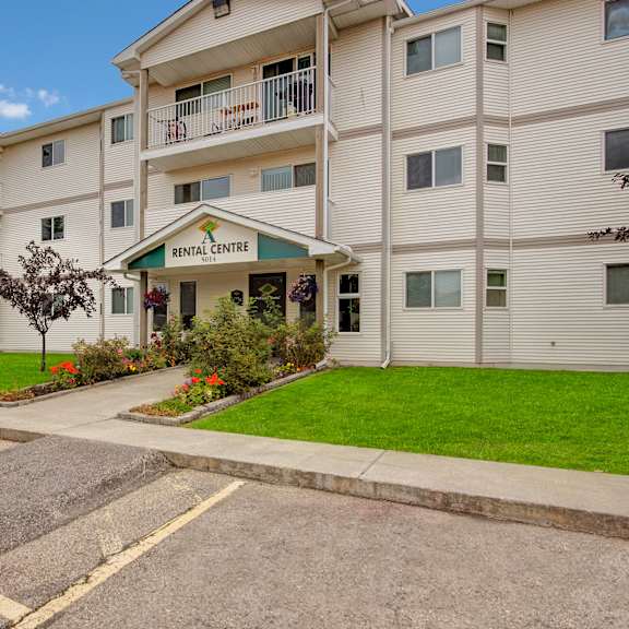 three-story beige birch terrace apartment homes apartment building with balconies, labeled "rental centre." surrounded by green lawn, trees, and colorful flowerbeds under a clear sky.