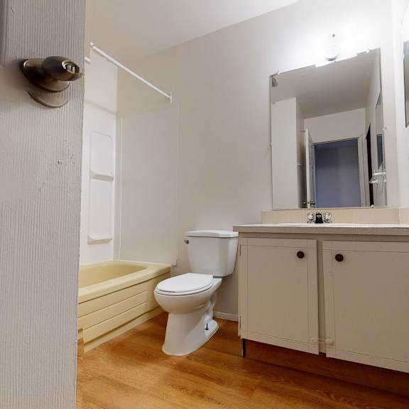 simple bathroom here at broadmoor apartment homes with a wooden floor, white vanity, and mirror. cream bathtub and toilet. light creates a clean, minimalistic feel.