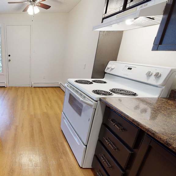 narrow kitchen here at broadmoor apartment homes with wooden floors, dark brown cabinets, and granite countertops. a white stove is on the right. glass door leads to a balcony.