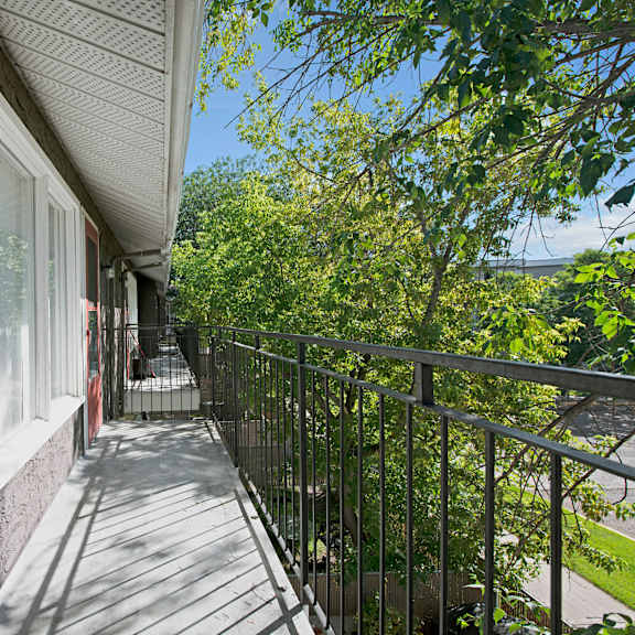 a narrow balcony here at broadmoor apartment homes with metal railing runs alongside a house with large windows. the balcony overlooks lush green trees on a sunny day, creating a serene atmosphere.