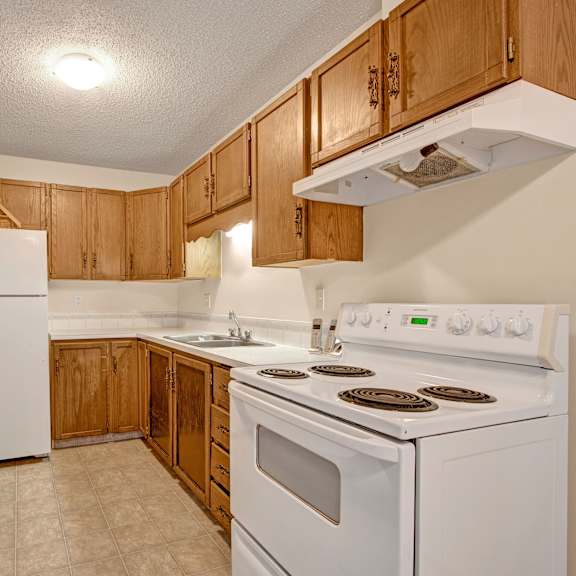 cozy kitchen here at broadmoor apartment homes with wooden cabinets, white countertops, and a tiled floor. features a white fridge, stove, and sink beneath warm lighting.