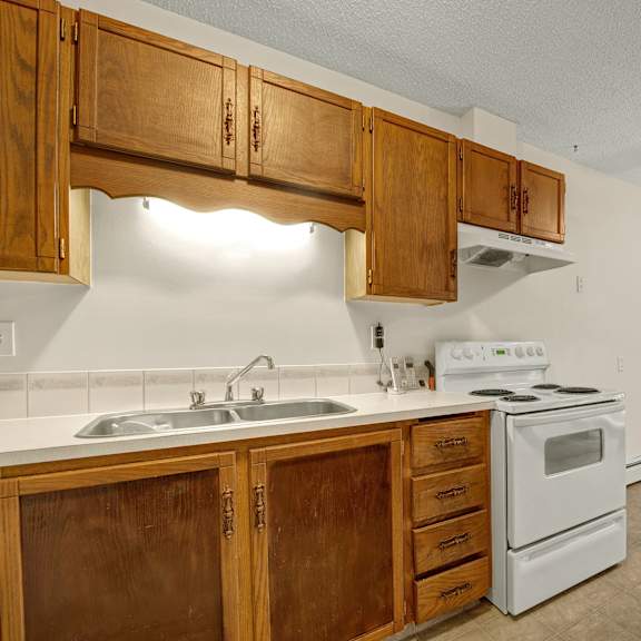 a cozy kitchen here at broadmoor apartment homes featuring wooden cabinets, a double sink, a white stove, and a ceiling fan. the space radiates warmth with its earthy tones.