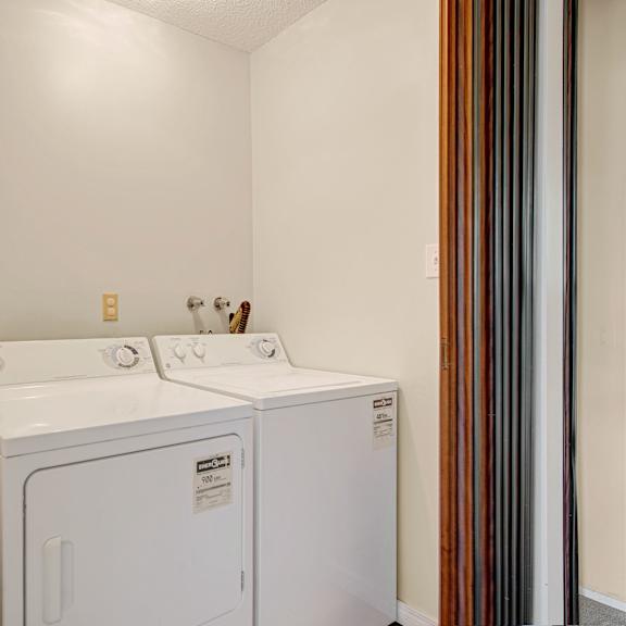 laundry room here at broadmoor apartment homes with a white washing machine and dryer side by side. to the left, a bathroom with a sink and mirror is partially visible. soft lighting fills the space.