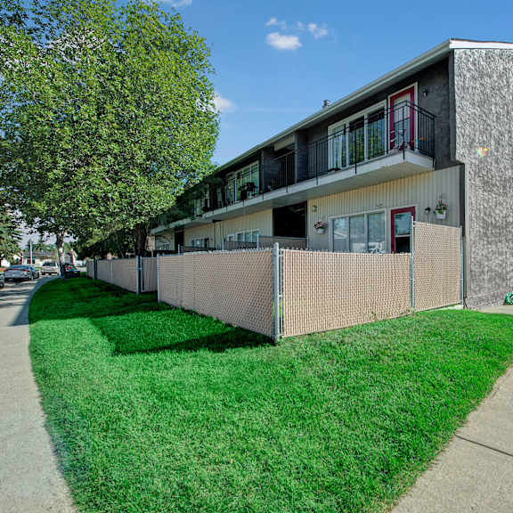 two-story gray broadmoor apartment homes building with balconies and chain-link fence, a lush green lawn, and a large tree. "now leasing" sign visible. bright, clear day.