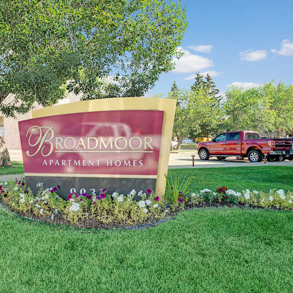 sign for broadmoor apartment homes in a landscaped area with colorful flowers and green grass. a red truck and trees are visible in the background, conveying a suburban setting.
