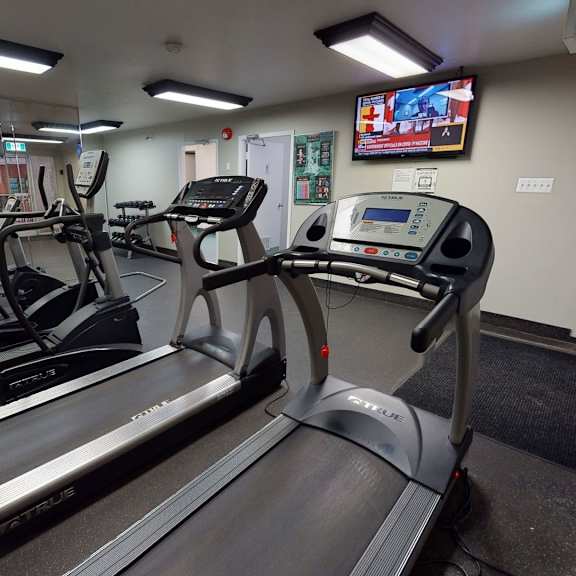 gym room here at cambridge west apartment homes with treadmills and exercise bikes facing a mirrored wall. a tv displays news in the background. the space is well-lit and organized.