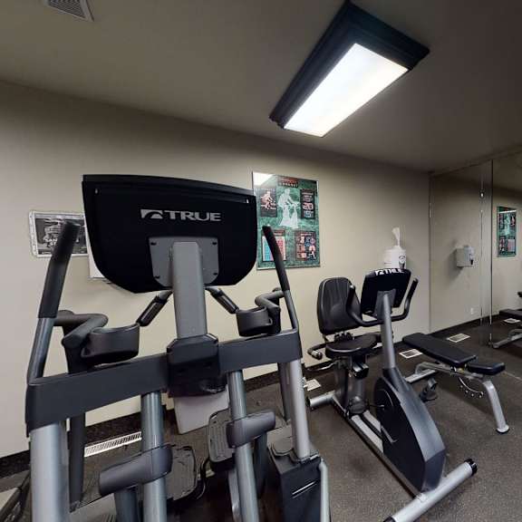 a small gym here at cambridge west apartment homes with exercise machines, including stationary bikes and elliptical trainers, facing a mirrored wall. posters are on the beige walls, creating a focused, quiet atmosphere.