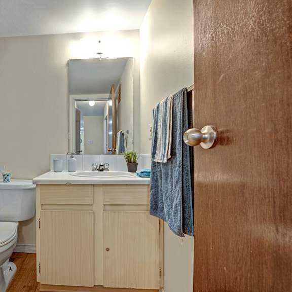 small bathroom here at cambridge west apartment homes with wooden door, floral shower curtain, toilet, and light wood vanity. a mirror above the sink reflects a cozy, simple decor.