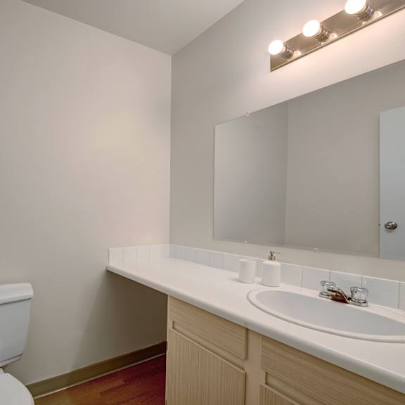 small, clean bathroom here at cambridge west apartment homes with beige walls, wooden vanity, white countertop, sink, mirror, and toilet. soft lighting and minimal decor create a simple, calm atmosphere.