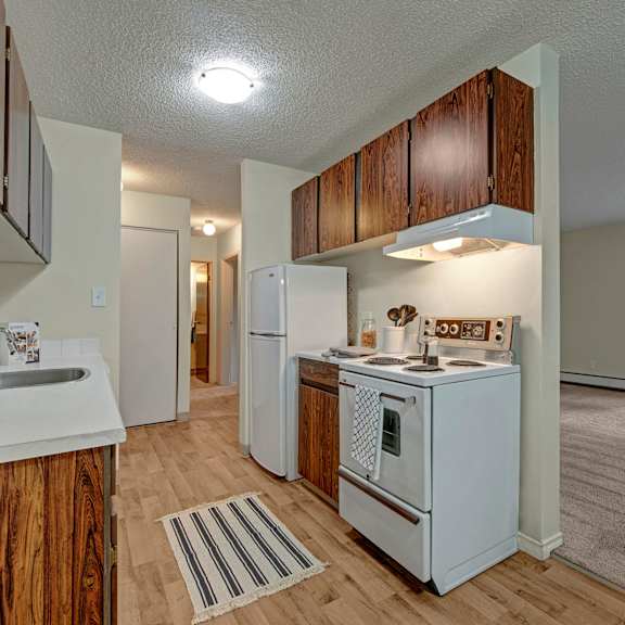 compact kitchen here at cambridge west apartment homes with wood cabinets, a white stove, and fridge on light wood flooring. bright and tidy, with an open view to a carpeted living area.