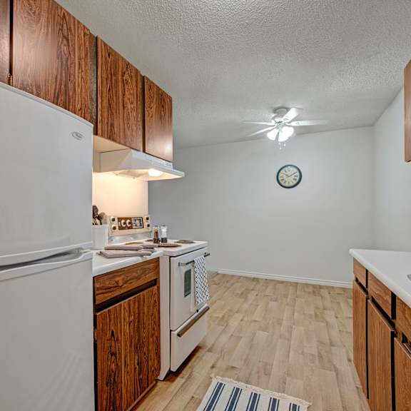 small kitchen here at cambridge west apartment homes with white appliances, wood cabinets, and light wood flooring. a ceiling fan and wall clock are visible. the space feels simple and cozy.
