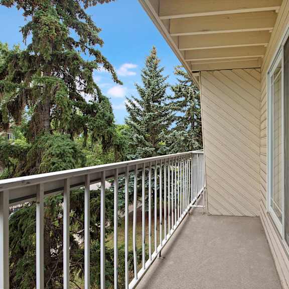 a cozy balcony here at cambridge west apartment homes with a beige railing and sliding glass doors, overlooking lush green pine trees and a clear blue sky, conveying a tranquil atmosphere.