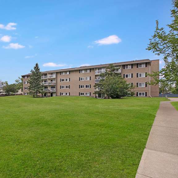 expansive green lawn with scattered evergreen trees leads to a four-story cambridge west apartment homes building under a clear blue sky, conveying tranquility and openness.
