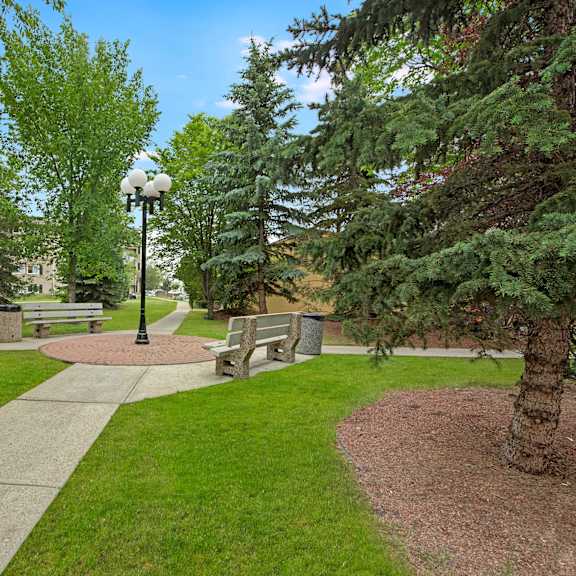 pathway in a tranquil park setting near cambridge west apartment homes with manicured lawns, benches, and a black lamp post. tall green trees create a serene atmosphere under a blue sky.