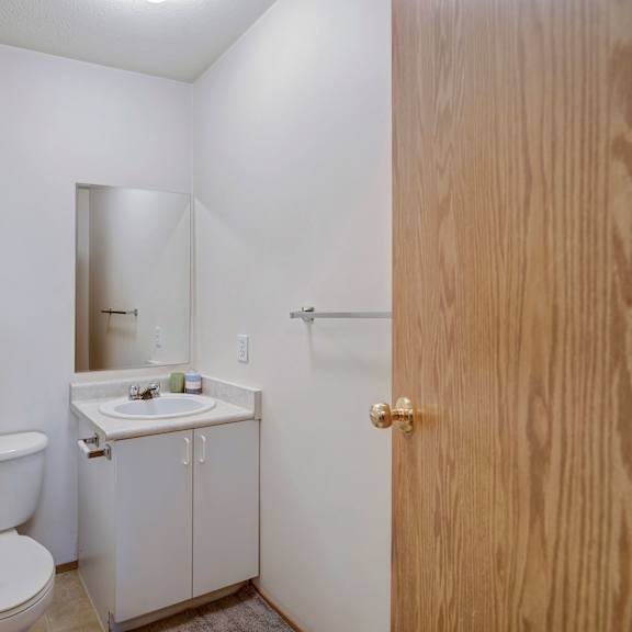 small, simple bathroom here at carrington place apartment homes with white walls and neutral tones. features a toilet, a small mirror above a sink, and wooden door, conveying a minimalistic tone.