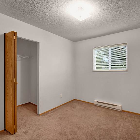empty bedroom here at carrington place apartment homes with beige carpet and light gray walls. open closet with a wooden door on the left, a small window with blinds on the right. cozy and minimalistic.