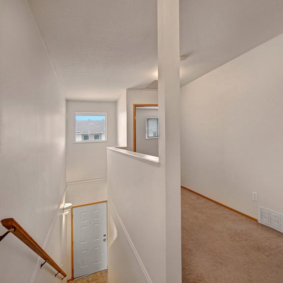 view of a simple, light-filled hallway here at carrington place apartment homes with white walls, a beige carpet, and a wooden railing on the stairs. a window with a view of rooftops is at the end, and a bathroom door with a towel is visible to the right.