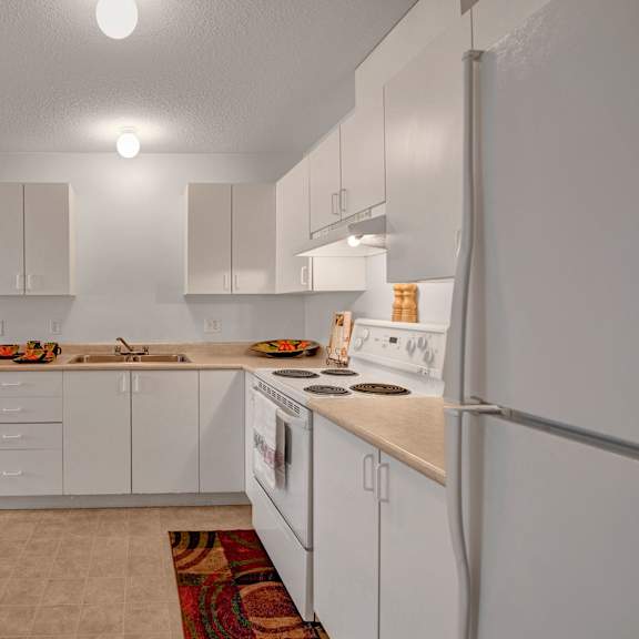 simple kitchen here at carrington place apartment homes with white cabinets, beige countertops, and white appliances, including a stove and fridge. a colorful rug and bowls add warmth.