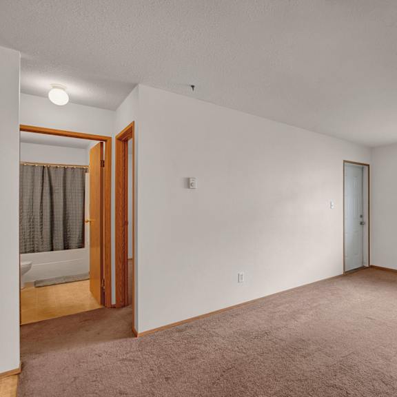 empty bedroom here at carrington place apartment homes with beige carpet and white walls. a window with blinds lets in soft daylight. open door reveals a bathroom with a shower curtain. minimalist tone.