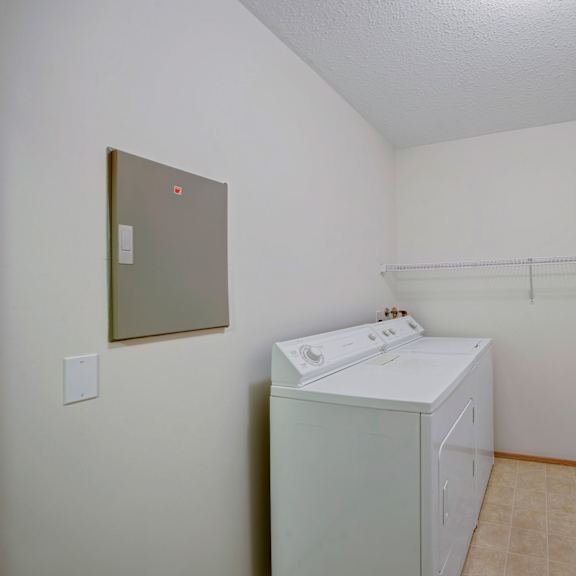 laundry room here at carrington place apartment homes with white washer and dryer, beige tiled floor, white walls, and a wire shelf above the appliances. clean, simple, and functional space.