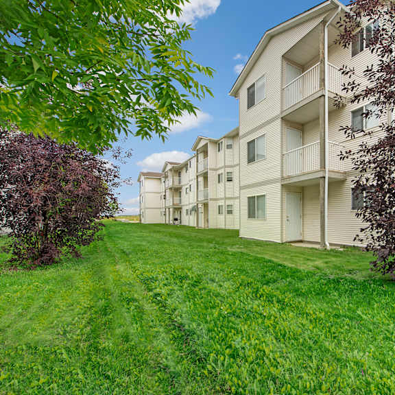 carrington place apartment homes buildings with beige siding line a lush, green lawn, bordered by trees with purple and green foliage against a blue sky with clouds.