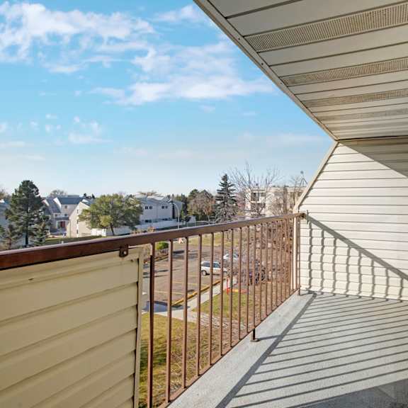 a sunlit balcony here at cedar gardens apartment homes with brown railings and sliding glass doors overlooks a scenic suburban neighborhood with trees and houses under a clear blue sky.