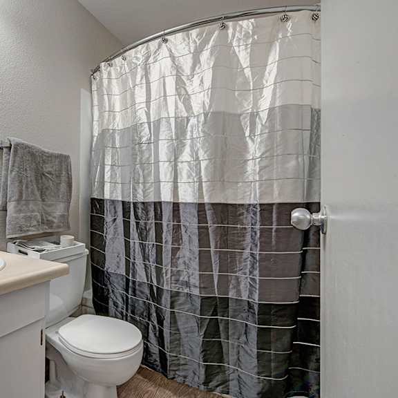 small bathroom here at cedar gardens apartment homes with a beige countertop, white sink, and silver faucet. gray towel hangs above the toilet. black-and-white shower curtain adds contrast.