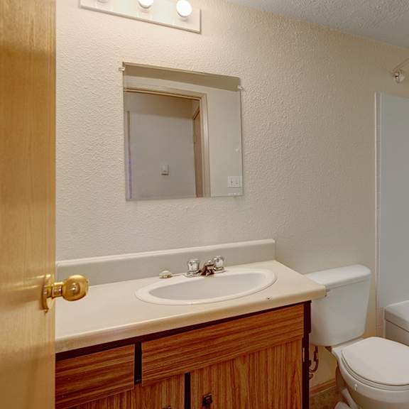 small, clean bathroom here at cedar gardens apartment homes with beige walls. features a wooden vanity, white sink, toilet, and bathtub with shower. bright lighting above the mirror adds warmth.