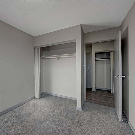 empty bedroom here at cedar gardens apartment homes with gray walls and carpet, featuring an open closet with hanging rods. doorway leads to hardwood floor hallway, creating a minimalist, neutral tone.
