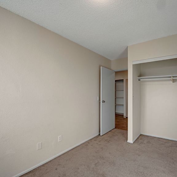 empty beige room here at cedar gardens apartment homes with a carpeted floor, partially open closet with sliding door, and an open door leading to a hallway with hardwood flooring. minimalist and neutral.