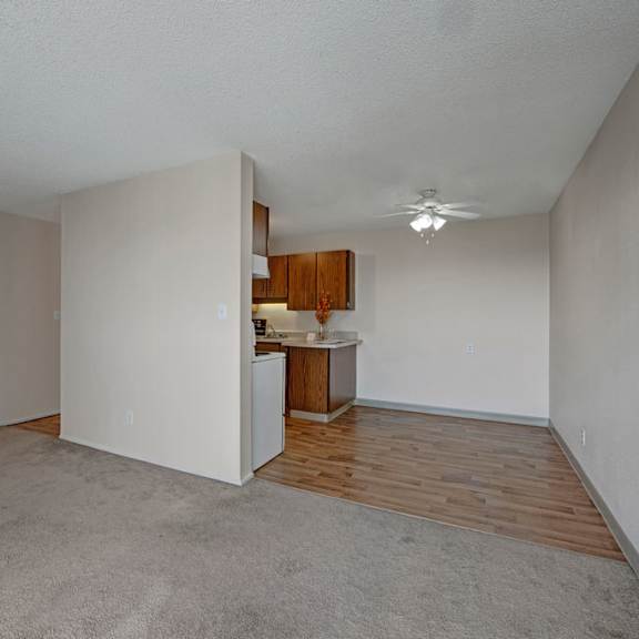 spacious, empty living room here at cedar gardens apartment homes with beige walls, carpeted floor, and a separate area with wood flooring. a small kitchen with wooden cabinets and ceiling fan.