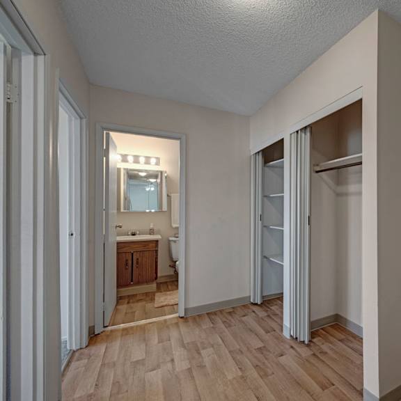 minimalist hallway here at cedar gardens apartment homes with light wood flooring. open closet with sliding doors on the right. bathroom with mirror and lights visible in the background.