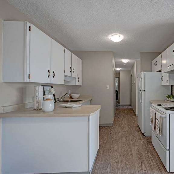 a modern, narrow kitchen here at cedar gardens apartment homes with white cabinets, an electric stove, and a fridge. light wood flooring and beige walls create a clean, neutral tone.